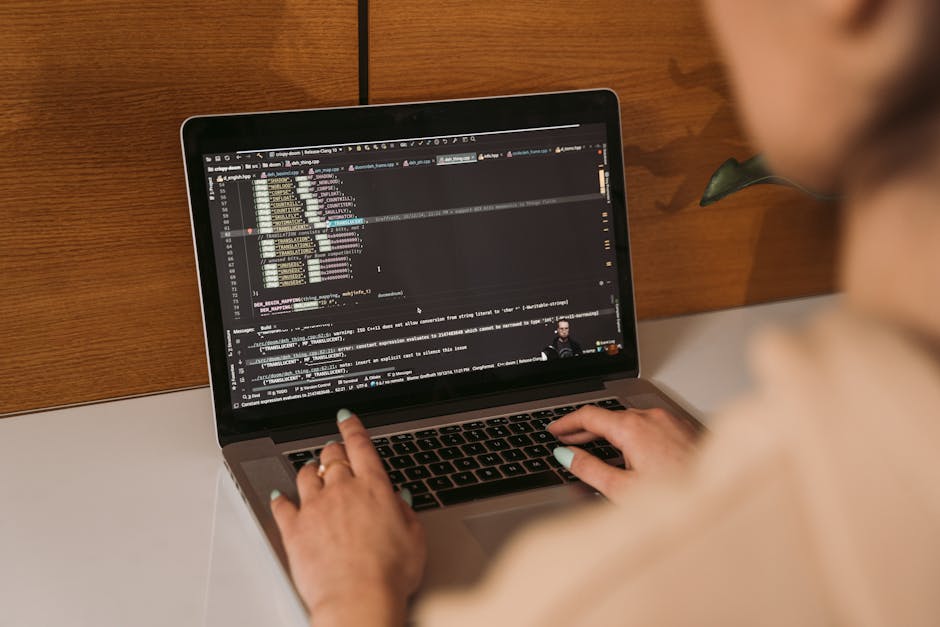 A close-up view of a laptop on a wooden desk displaying lines of code on the screen, with a partially visible orange mug with white text and a white notebook with a red border placed beside it, illumi